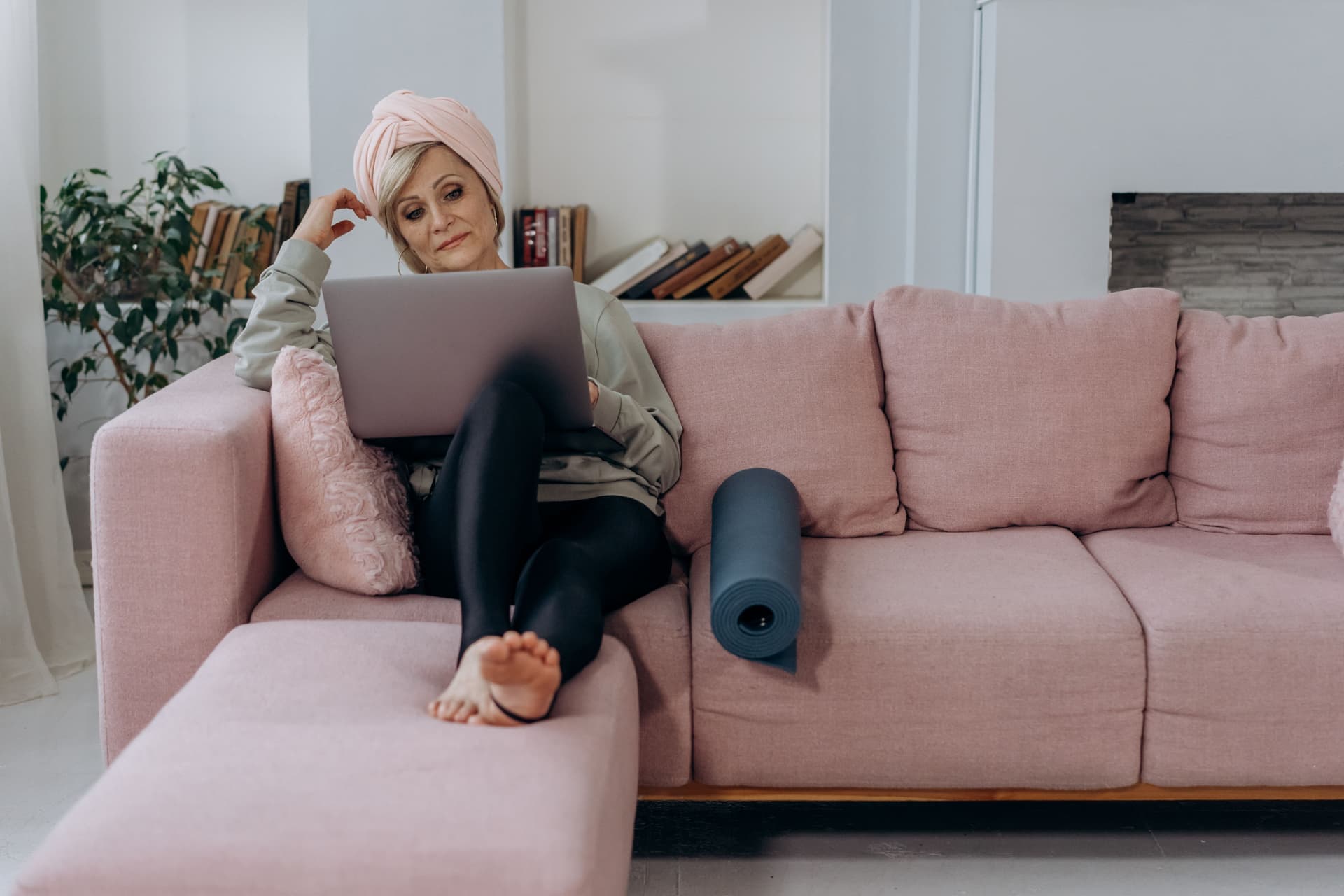 Woman on a couch with a laptop and a rolled yoga mat, ready for a home workout